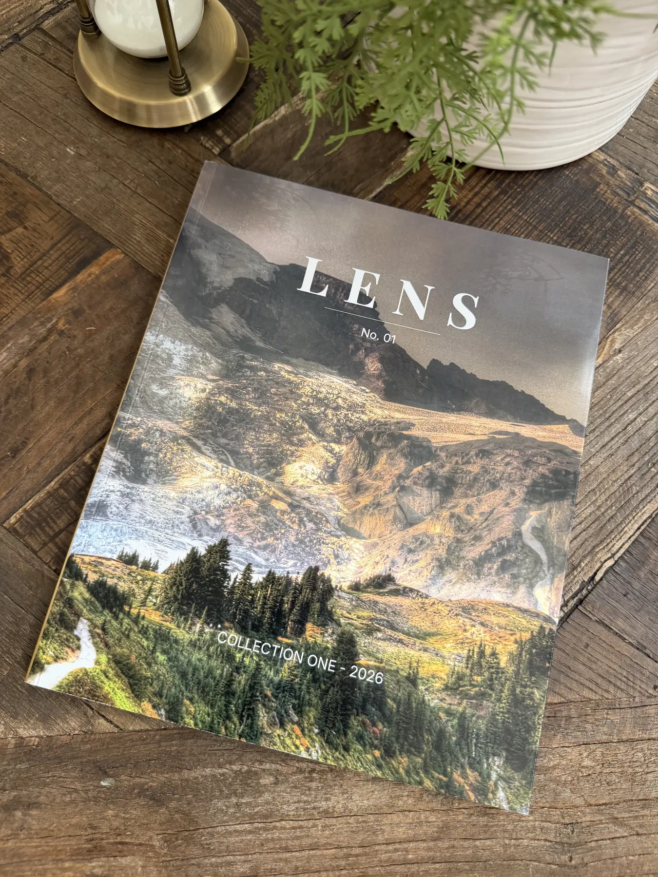 A copy of LENS lying closed on a wooden coffee table beside a brass lamp and a potted fern, daylight falling across the cover.