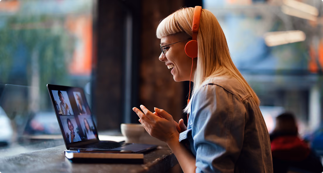 Woman with orange headphones smiling and video chatting on laptop in a cozy cafe.