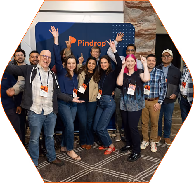 A diverse group of people posing cheerfully in front of a blue backdrop with the orange Pindrop logo, some raising their hands and smiling.