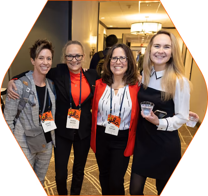 Four women standing together in a hallway at an event, smiling and wearing name badges and event lanyards; one woman on the left wears a gray checkered suit with a backpack, the second wears a black jacket and red top, the third wears a red jacket over a white top with black stripes, and the fourth wears a black dress with white collar and cuffs holding a clear plastic cup.