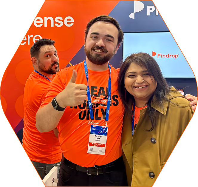 Three smiling individuals at a booth wearing orange shirts, one man in front giving a thumbs up, the woman on the right wearing a tan coat, with a Pindrop logo displayed behind them.