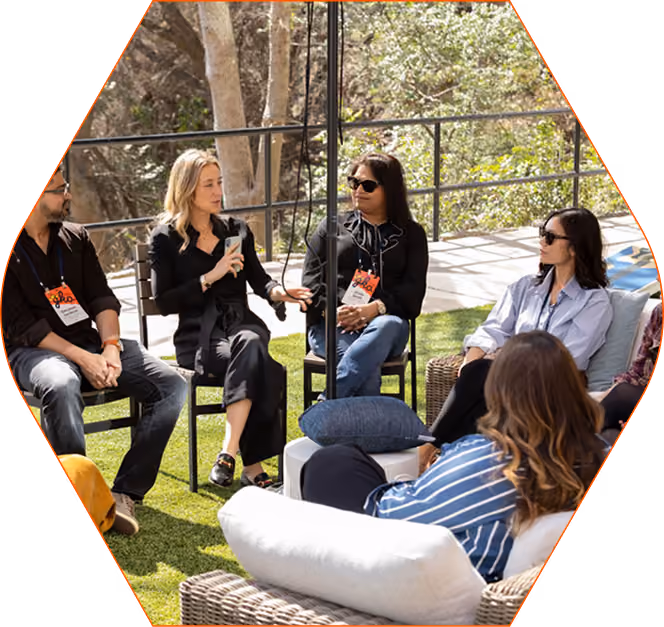 Group of five adults seated in a semi-circle outdoors on a green lawn, engaged in a discussion, with some holding microphones and others listening attentively.