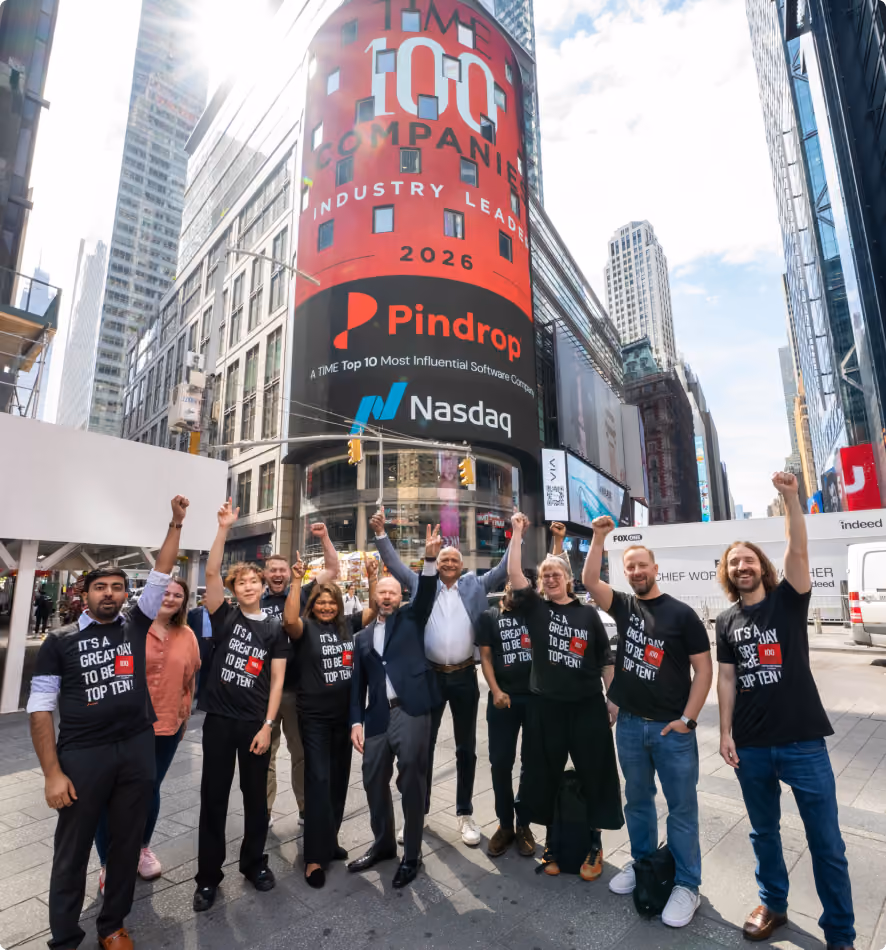Group of people standing in Times Square, New York City, raising fists and smiling in front of a large vertical digital billboard that reads 'TIME 1000 Companies Industry Leaders 2026' with logos for Pindrop and Nasdaq beneath it.