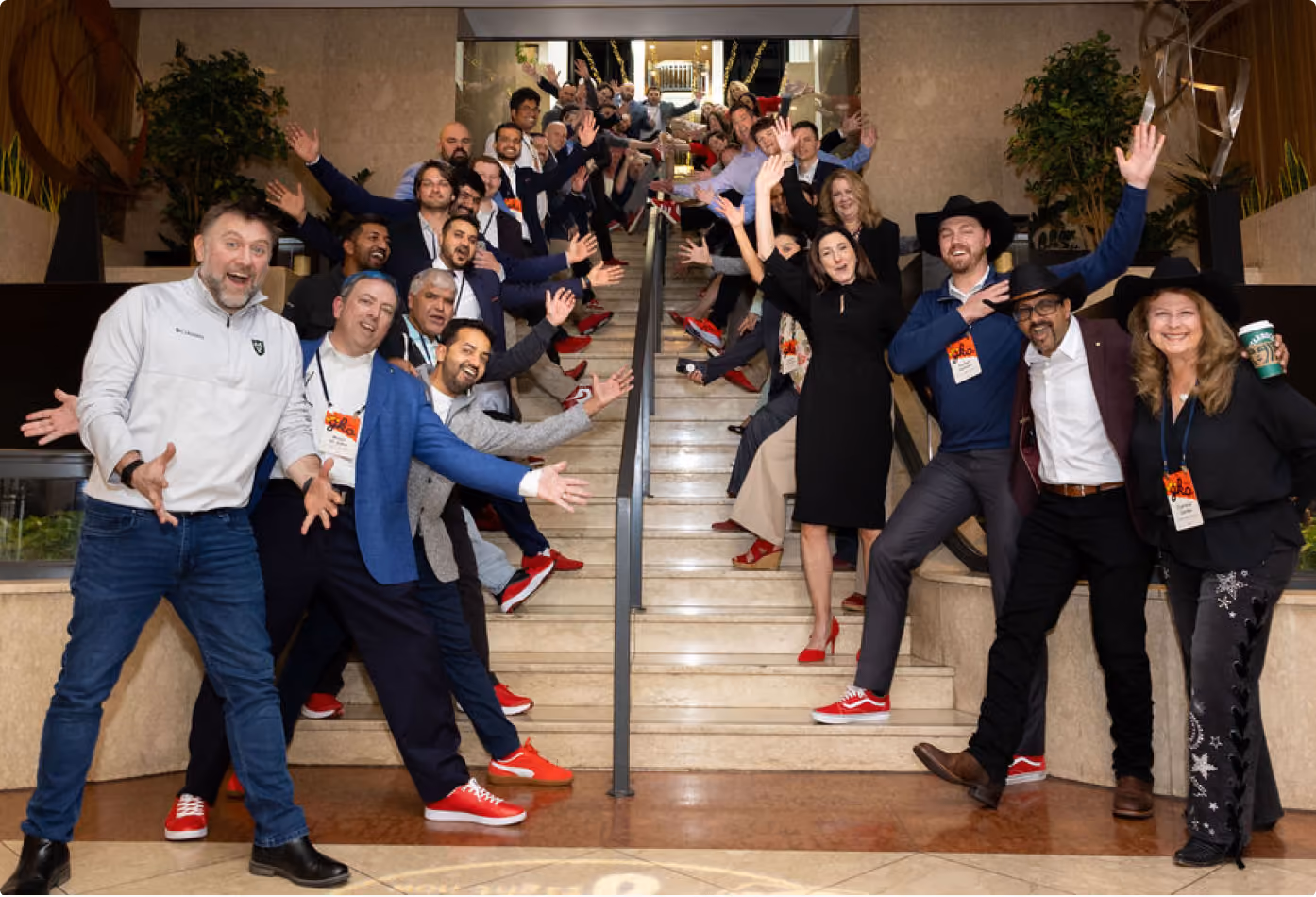 Large group of diverse people posing cheerfully on a wide staircase, many wearing red shoes and waving hands, some wearing hats, in a well-lit indoor space.