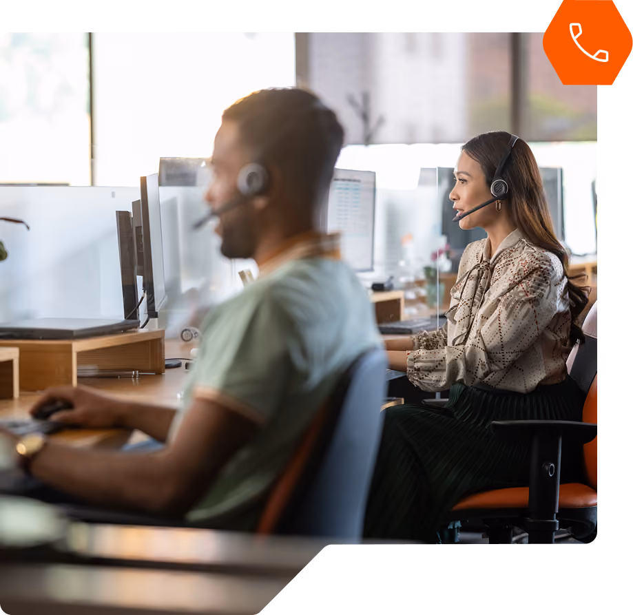 Two call center representatives with headsets looking at computers sitting at desks in office side by side. icon of phone layered on top in orange hexagon