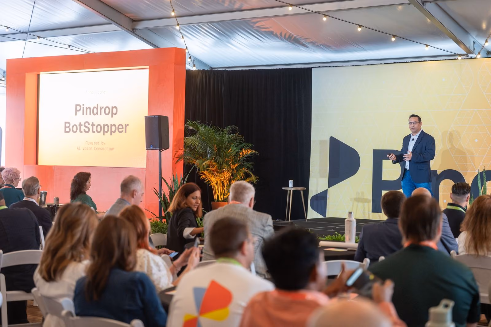 SVP of Product at Pindrop, Amit Gupta, speaks in front of a crowd at Pindrop's 2026 CFX Summit. A screen to the left shows an announcement for Pindrop BotStopper.