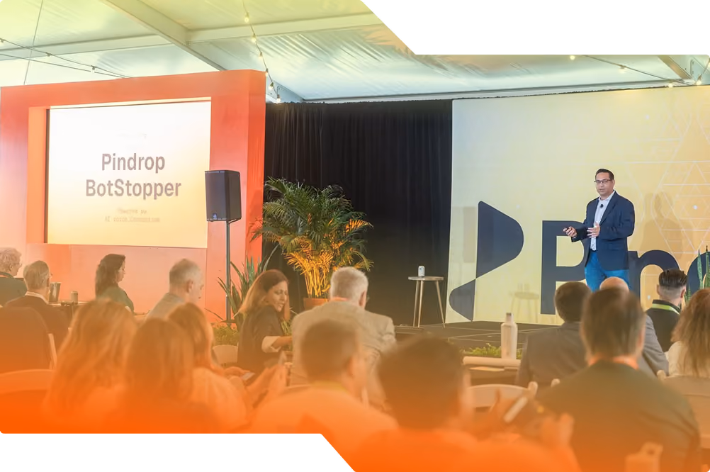 SVP of Product at Pindrop, Amit Gupta, speaks in front of a crowd at Pindrop's 2026 CFX Summit. A screen to the left shows an announcement for Pindrop BotStopper.