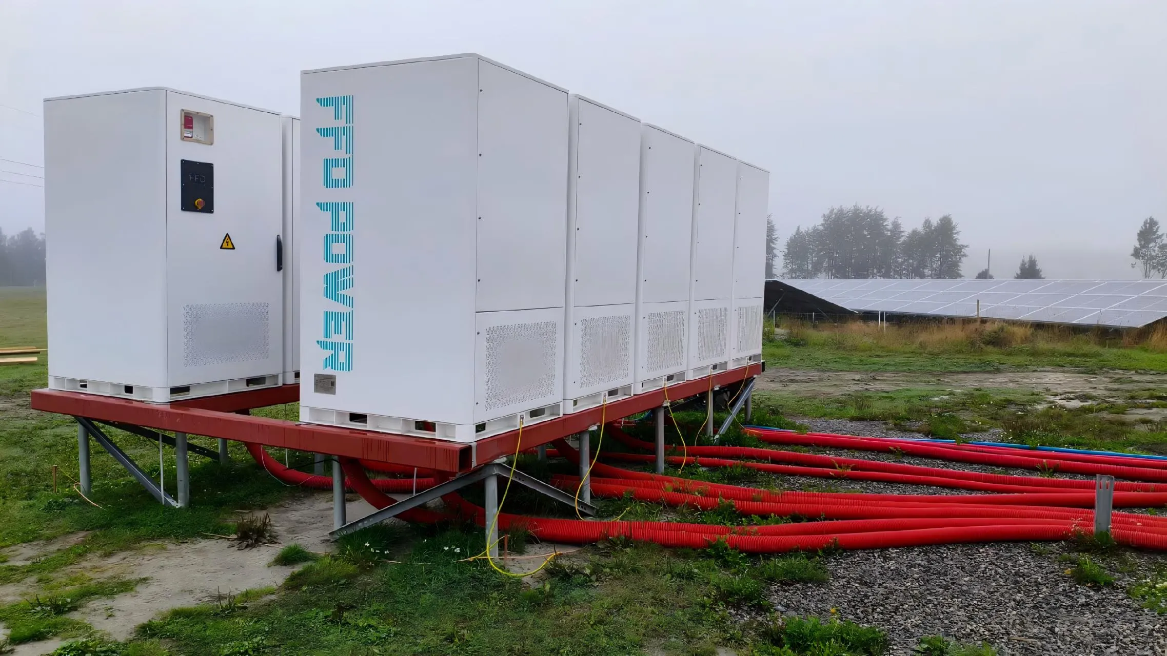 FFD POWER Batterieschränke auf einer erhöhten Stahlplattform in nebliger Wiesenlandschaft, mit Solarpark im Hintergrund und roten Kabelschutzrohren