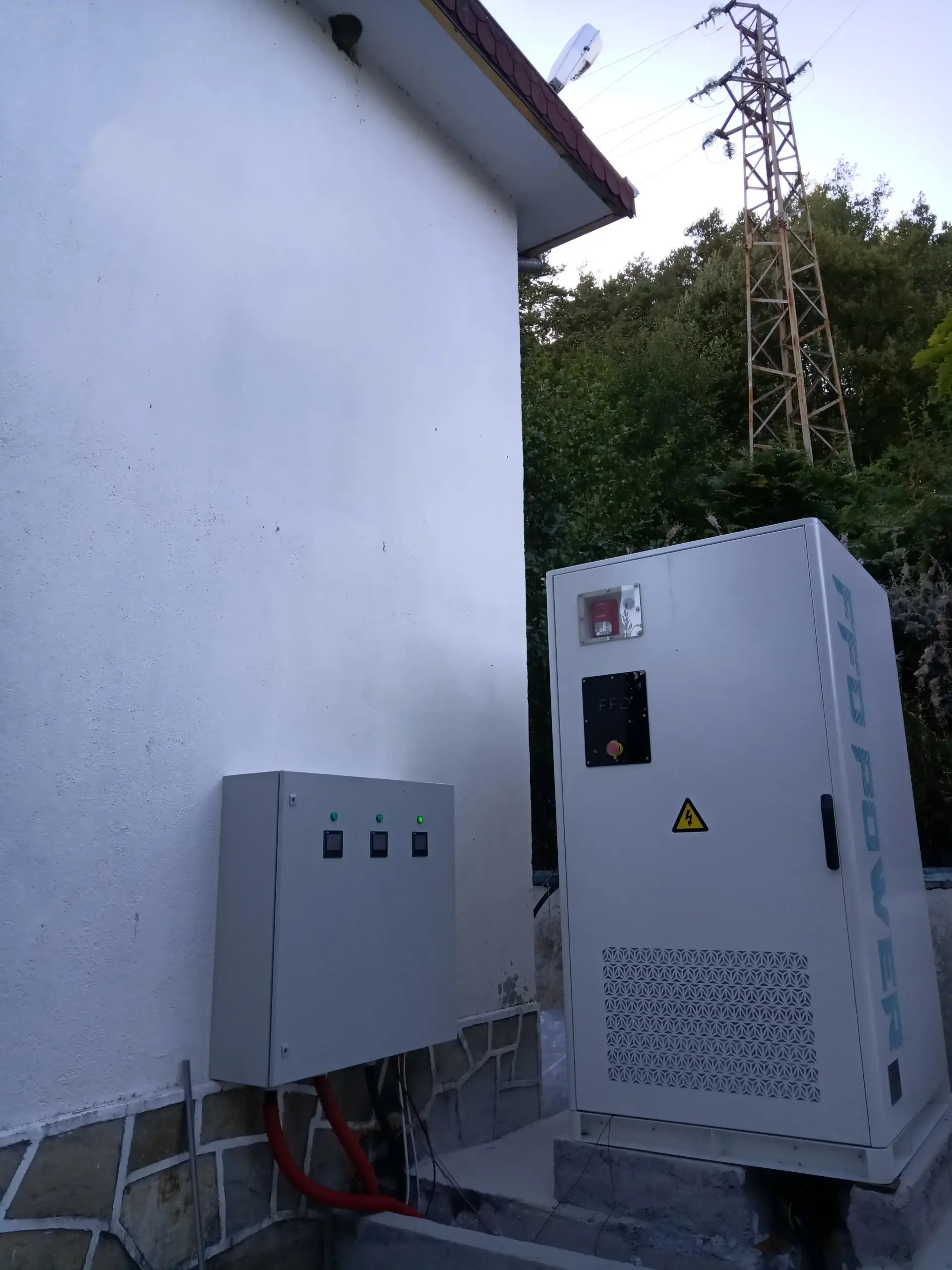 FFD POWER Batterieschrank neben einem Wohnhaus, im Hintergrund ein Hochspannungsmast