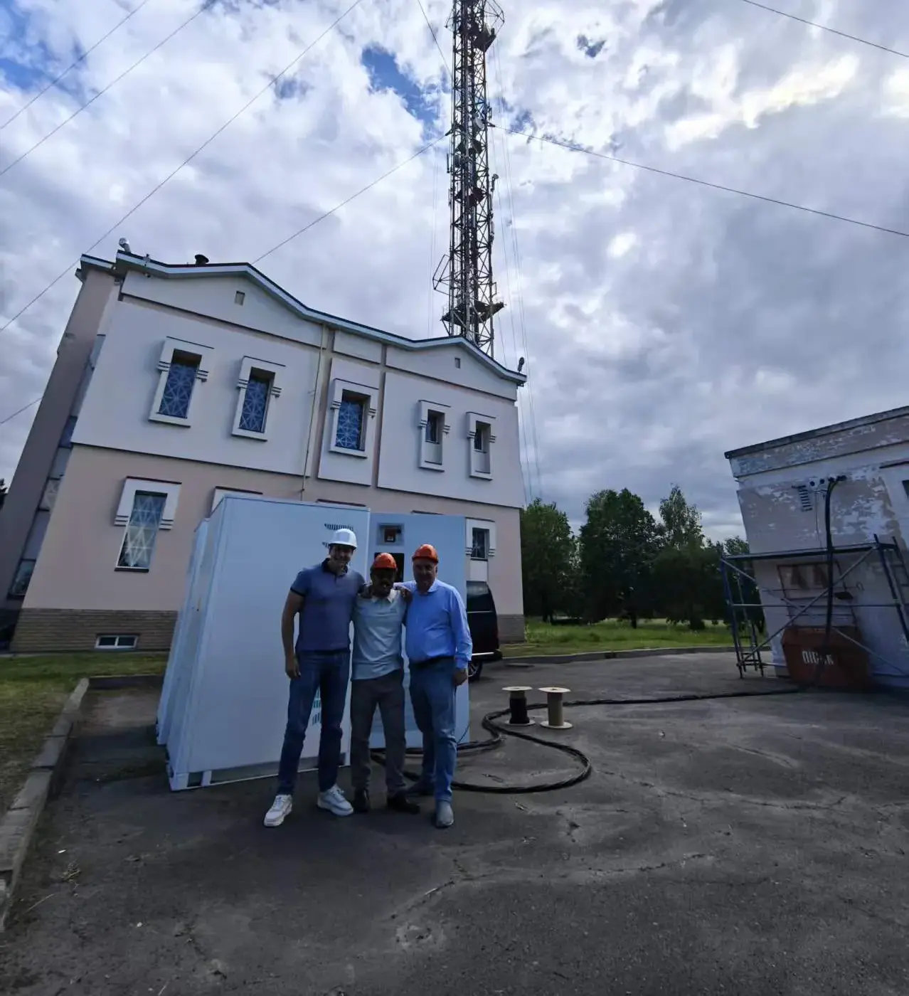 Drei Personen mit Schutzhelmen vor einem FFD POWER Batterieschrank, im Hintergrund ein Sendemast und Kabeltrommeln