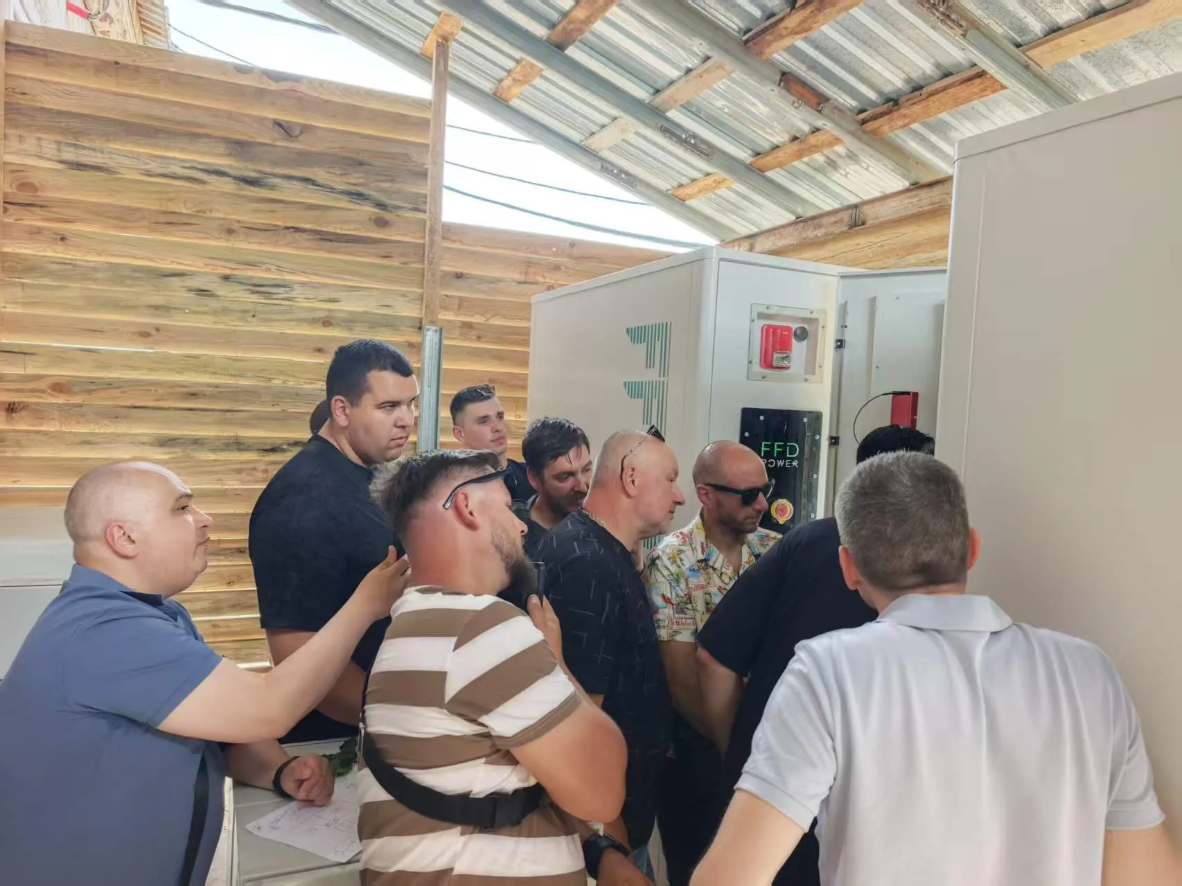 Eine Gruppe von Personen versammelt sich um einen FFD POWER Batterieschrank unter einem provisorischen Holzdach bei einer Standortbesichtigung