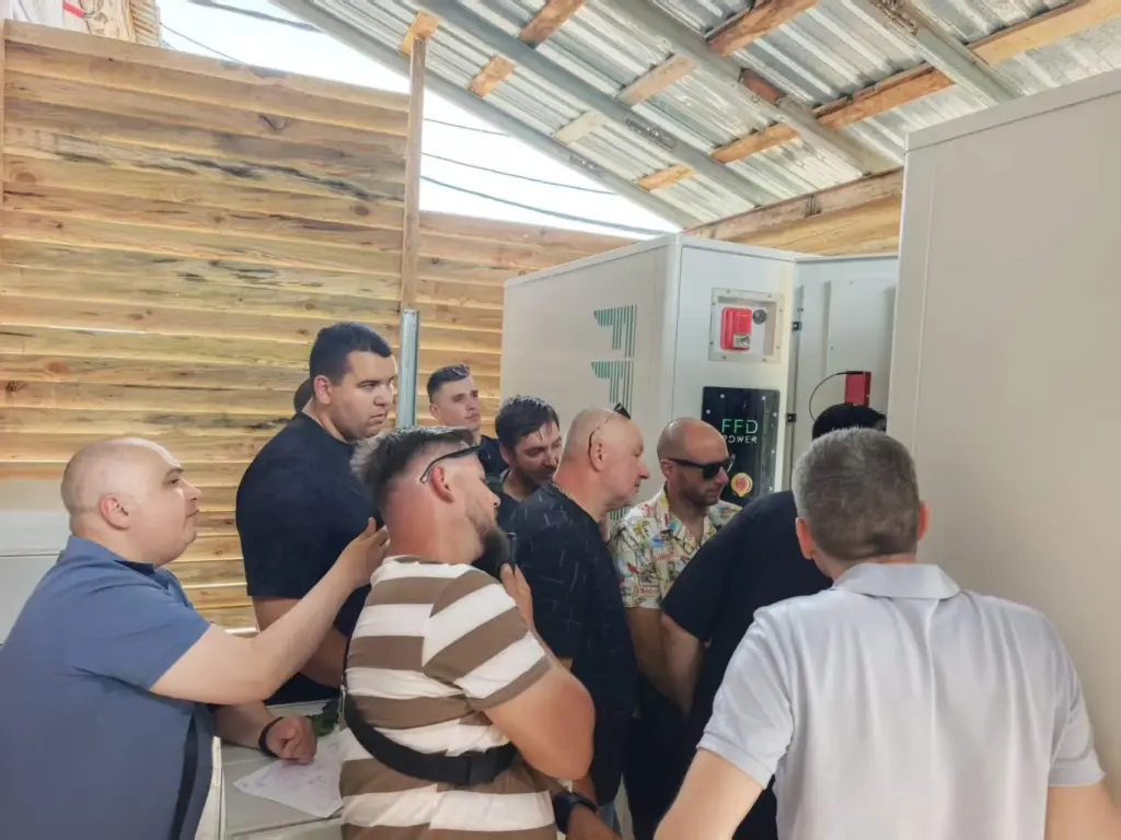 Eine Gruppe von Personen versammelt sich um einen FFD POWER Batterieschrank unter einem provisorischen Holzdach bei einer Standortbesichtigung