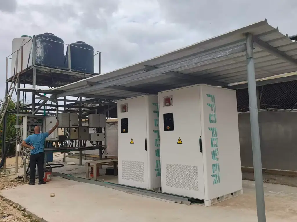 Zwei FFD POWER Batterieschränke unter einem Metalldach neben einem Wasserturm und Steuerungspaneelen, ein Mitarbeiter prüft die Anlage