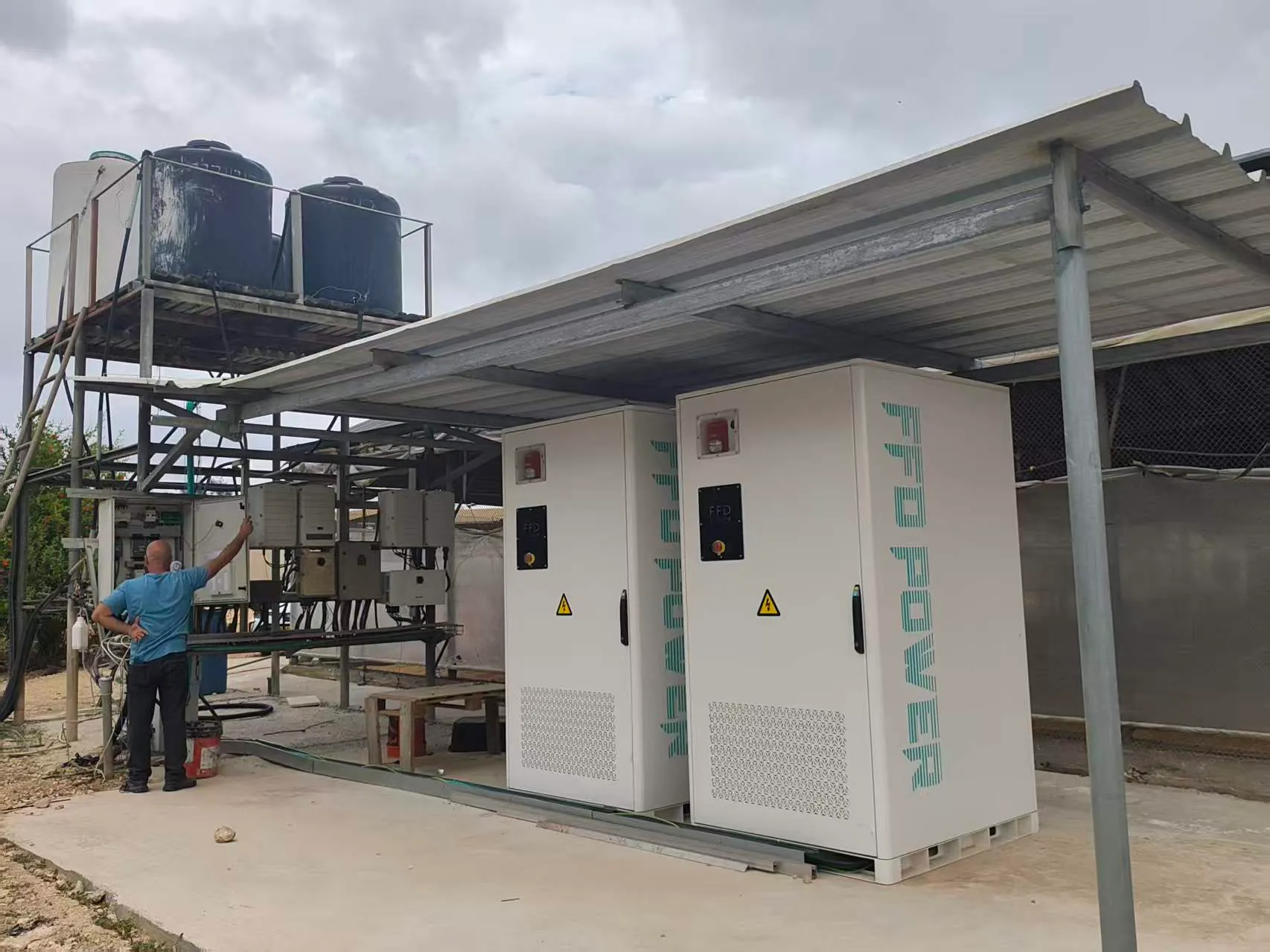 Zwei FFD POWER Batterieschränke unter einem Metalldach neben einem Wasserturm und Steuerungspaneelen, ein Mitarbeiter prüft die Anlage