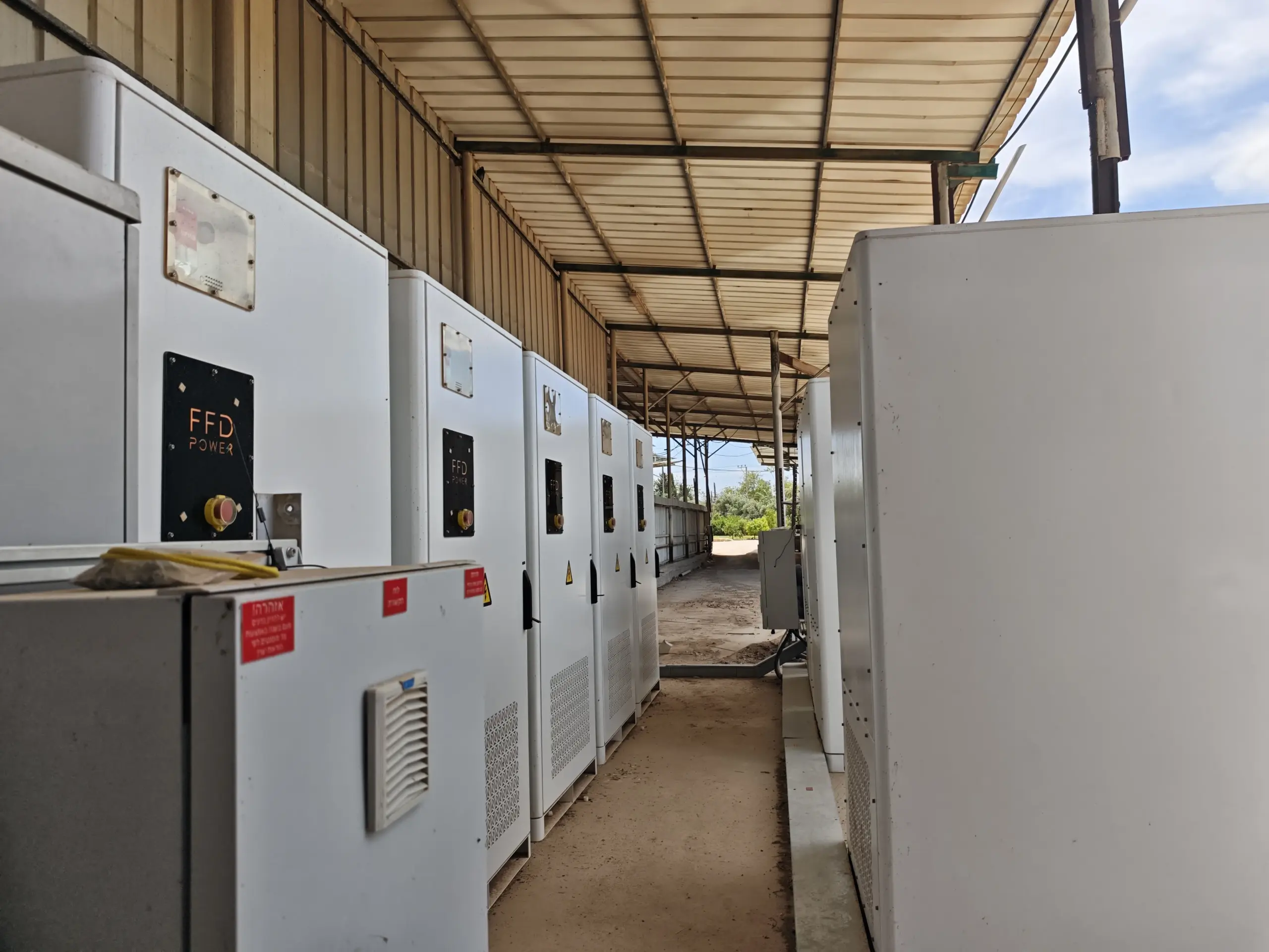 FFD POWER Batterieschränke beidseitig eines schmalen Gangs unter einem überdachten Bereich, mit hebräischen Warnhinweisen