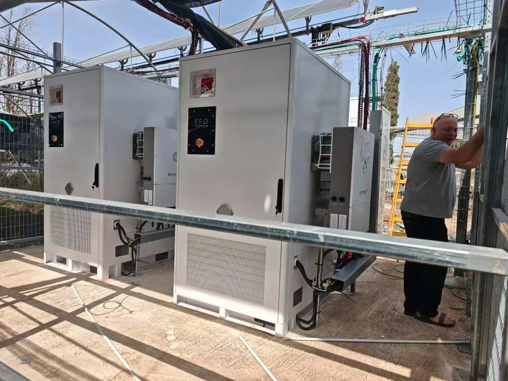 Zwei FFD POWER Batterieschränke mit seitlich montierten Wechselrichtermodulen in einem Gewächshausbereich, ein Arbeiter installiert die Umzäunung