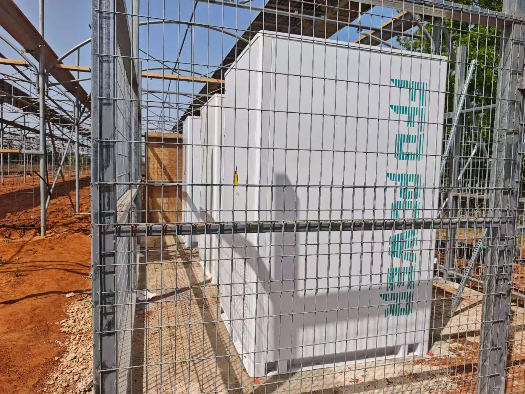 FFD POWER Batterieschrank in einem umzäunten Bereich auf rötlichem Boden, im Hintergrund eine im Bau befindliche Gewächshauskonstruktion