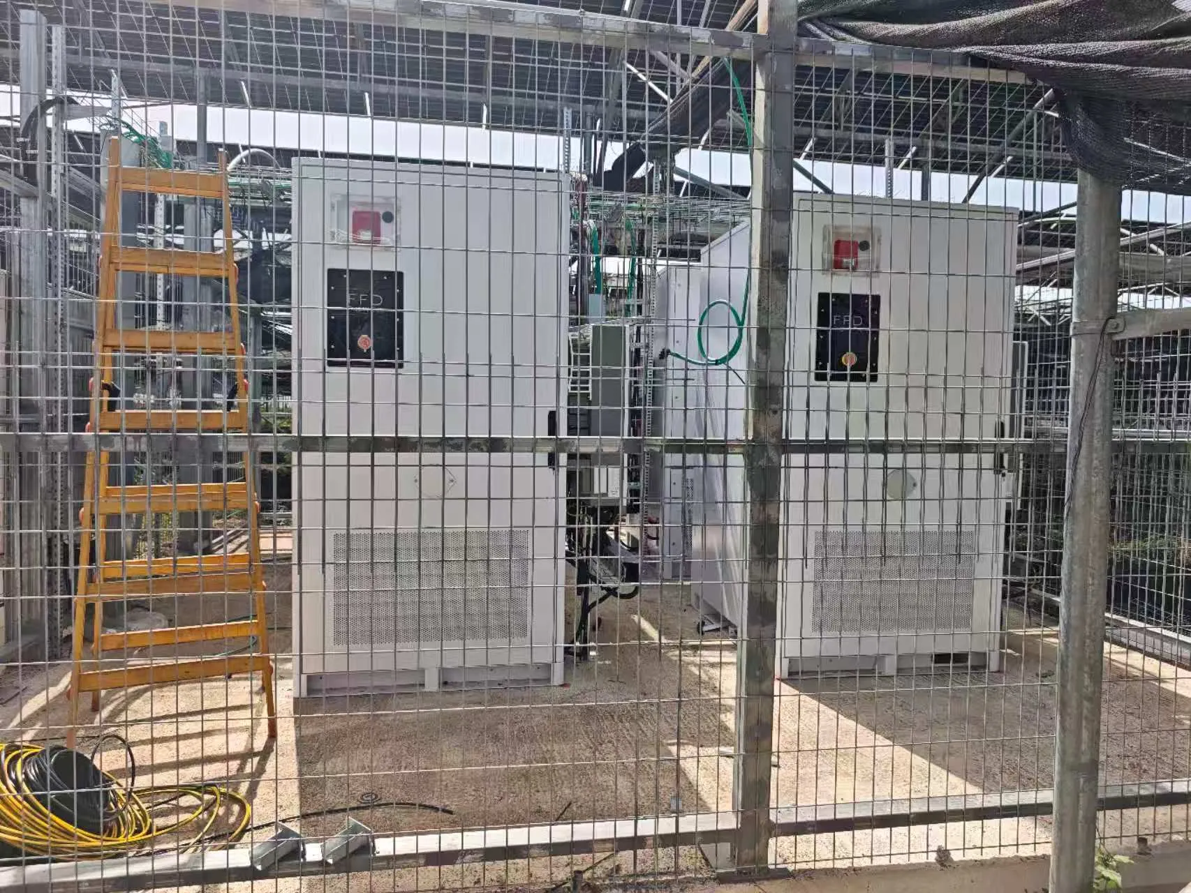 FFD POWER Batterieschränke in einem umzäunten Bereich unter einer PV-Gewächshauskonstruktion, mit Werkzeug und Kabeln am Boden