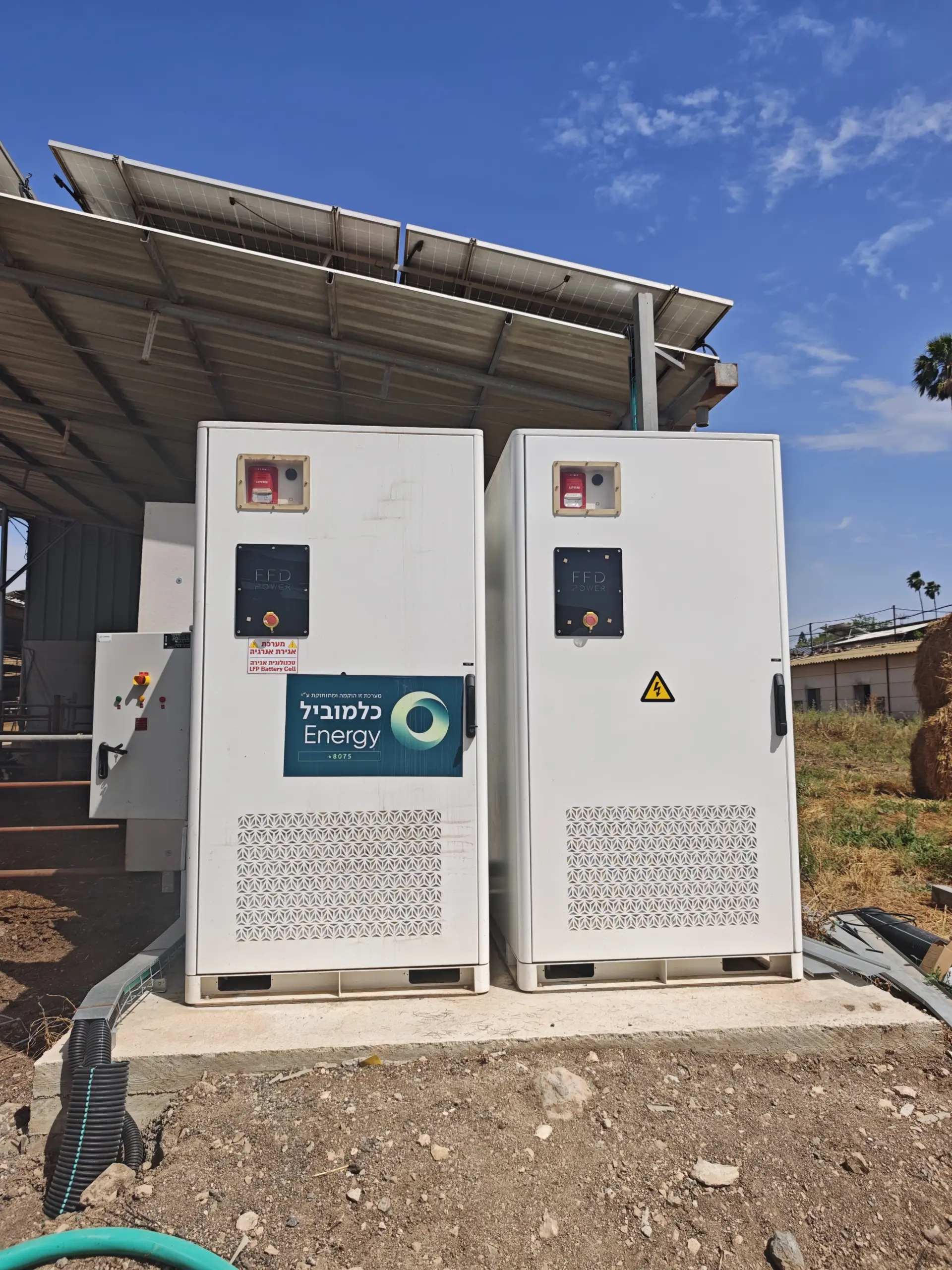 Zwei FFD POWER Batterieschränke unter einer Gewächshaus-PV-Konstruktion in Frontalansicht, mit Partner-Aufkleber „Kalmubil Energy" und Strohballen im Hintergrund