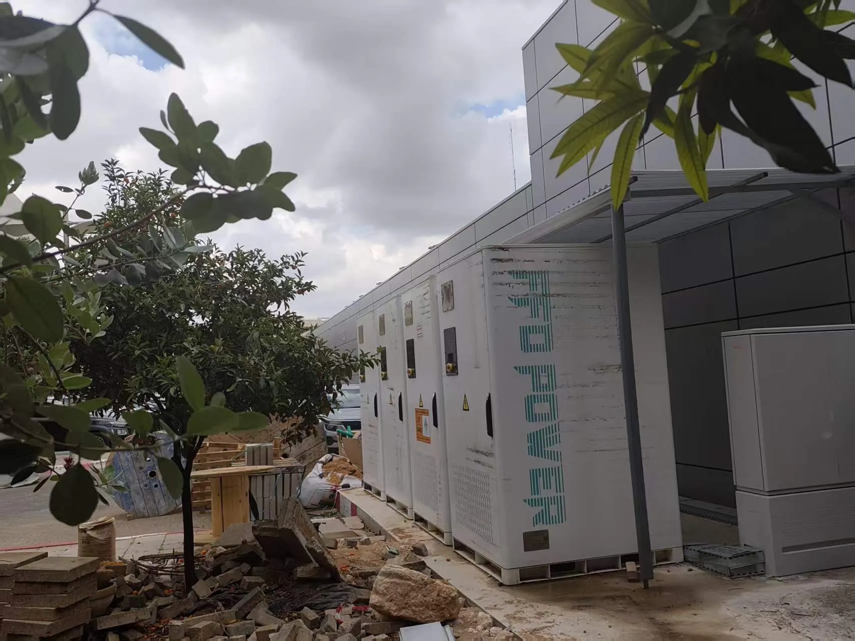 FFD POWER Batterieschränke entlang einer Gewerbegebäude-Fassade an einer Baustelle, im Vordergrund Bäume und Baumaterial