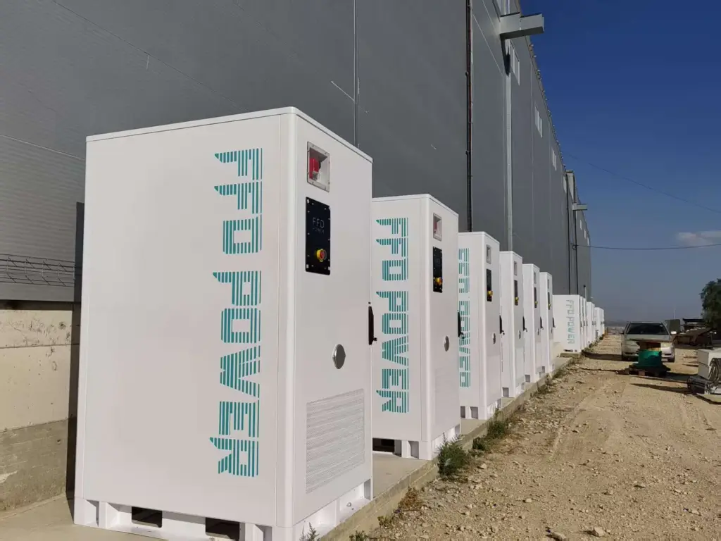 FFD POWER Batterieschränke entlang einer Industriehallen-Außenwand auf unbefestigtem Gelände