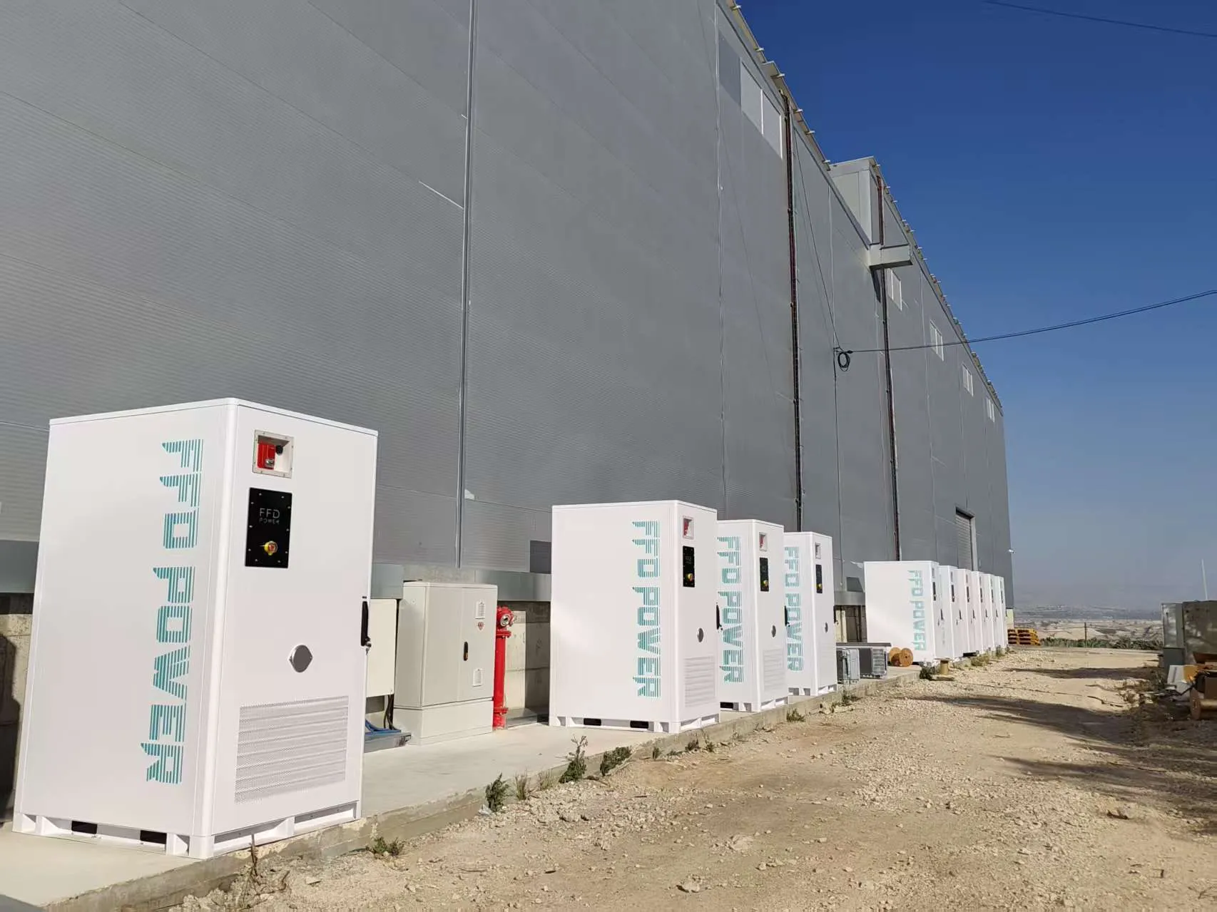 FFD POWER Batterieschränke entlang einer großen Industriehallen-Fassade, mit rotem Hydranten und weiteren Schränken in der Ferne