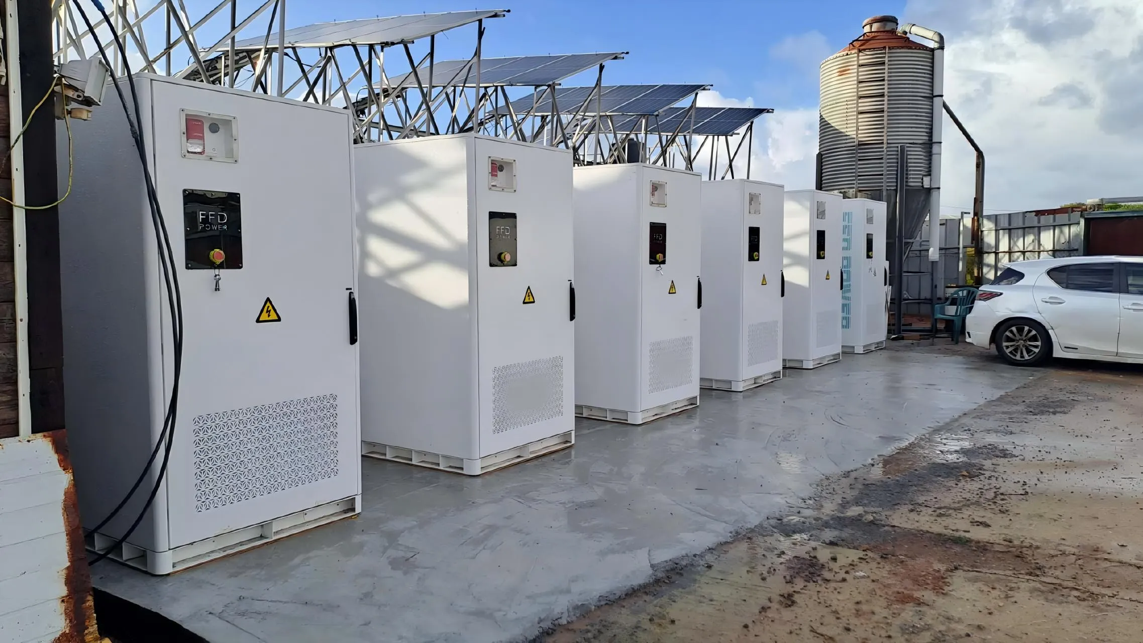 Reihe von FFD POWER Batterieschränken auf einem landwirtschaftlichen Betriebsgelände unter einer erhöhten PV-Anlage, mit Getreidesilo im Hintergrund