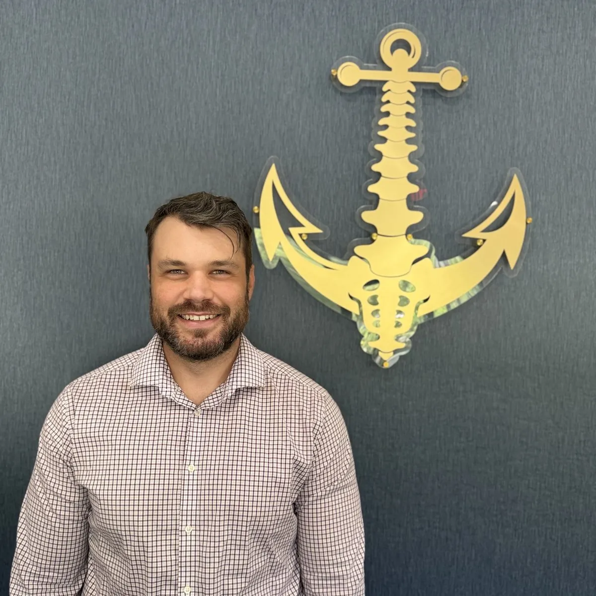 Dr. Ryan, chiropractor at Treasure Coast Spine & Sport, smiling in front of the practice's anchor-and-spine logo