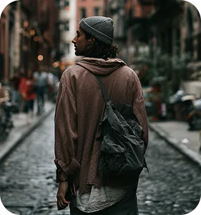 Person wearing a beanie and backpack walking down a cobblestone city street, seen from behind.