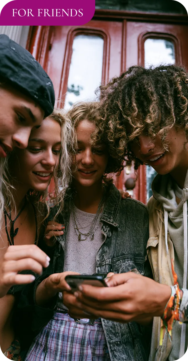 Four young people gathered closely, smiling and looking at a smartphone together outdoors.