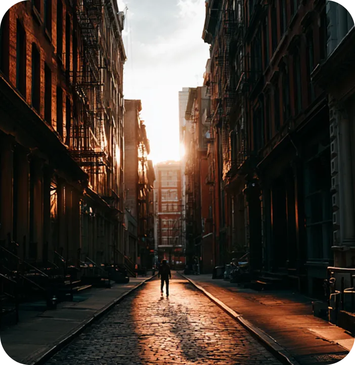 Person walking on a sunlit cobblestone street lined with tall buildings casting shadows.