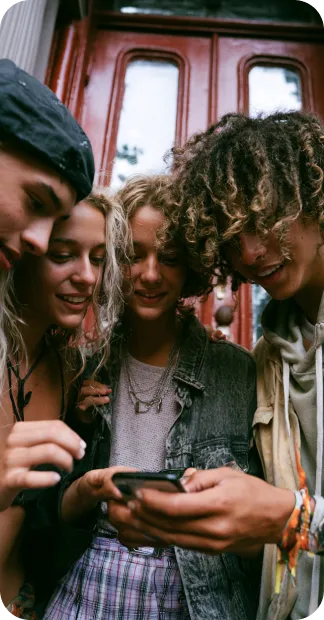 Four diverse young adults closely looking at a smartphone together outside a building.