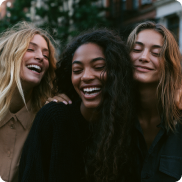 Three women smiling and embracing each other outdoors with blurred background.