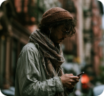 Person wearing a beanie and scarf looking down at their phone on a city street.