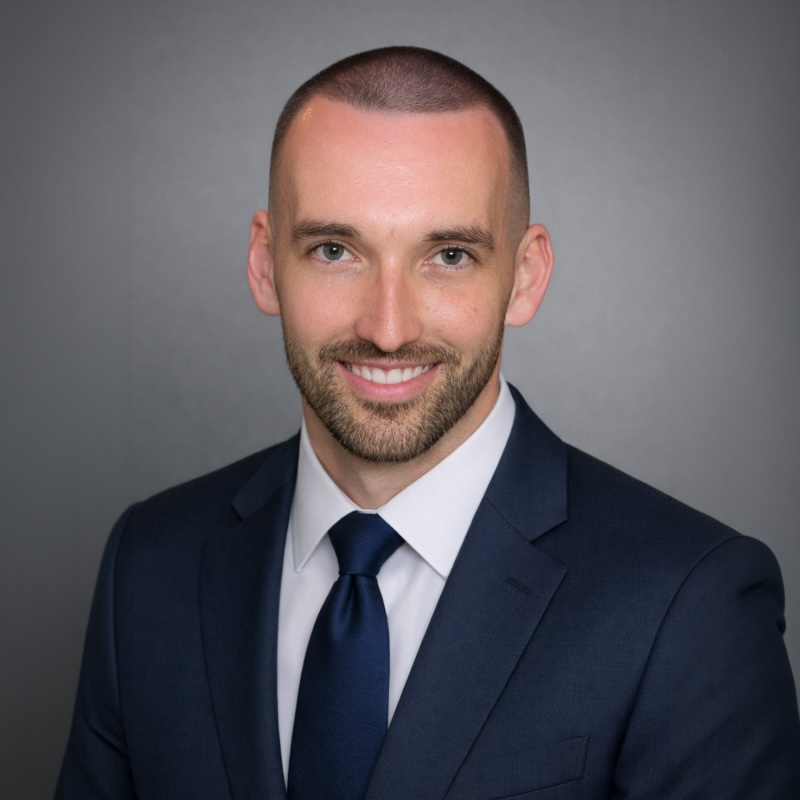 Smiling man with short hair and beard wearing navy suit, white shirt, and navy tie on gray background.