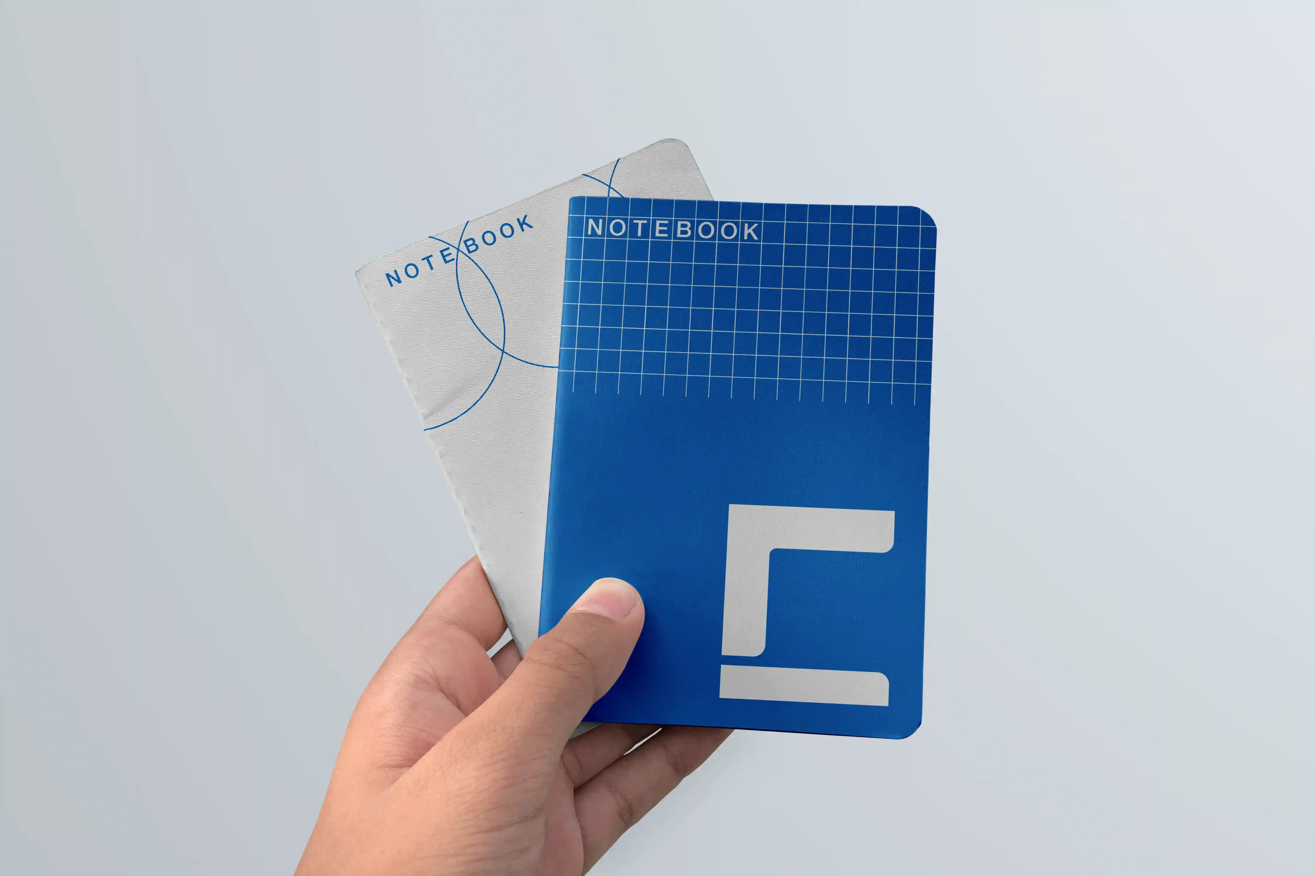 Hand holding two small notebooks, one blue with grid pattern and one light gray with circle lines.