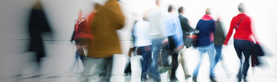 Blurred, motion-streaked image of a crowd of people walking through a light-filled indoor space, suggesting movement and pace.