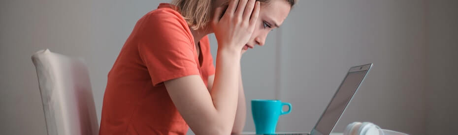 Woman in an orange t-shirt sitting at a table with her head resting on her hand, looking stressed while facing a laptop, with a teal mug beside her.