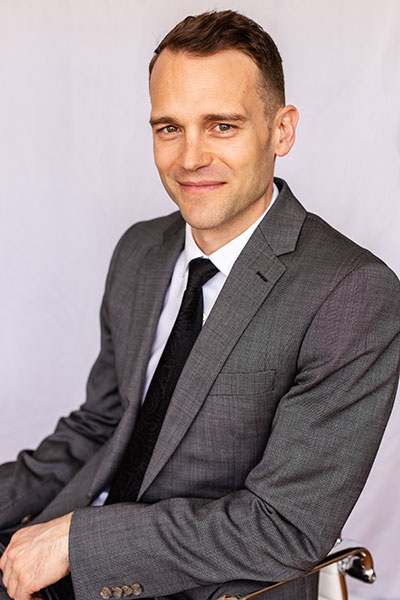 Cody Wagner wearing a grey suit with a black tie sitting in a chair