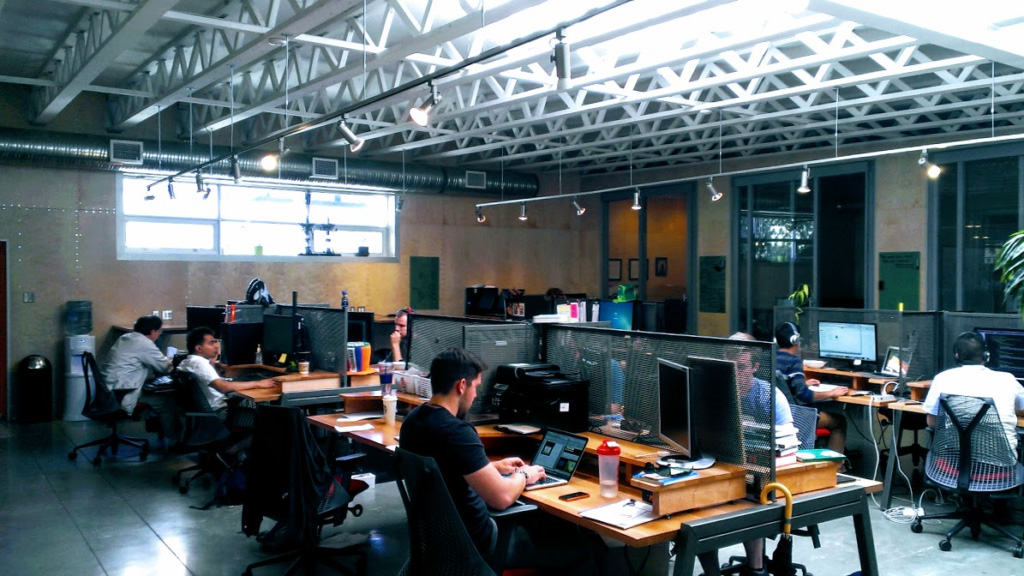Wide view of an open-plan office with an industrial exposed-truss ceiling, several employees seated at desks working on laptops and desktop monitors, with mesh cubicle dividers and large windows in the background.