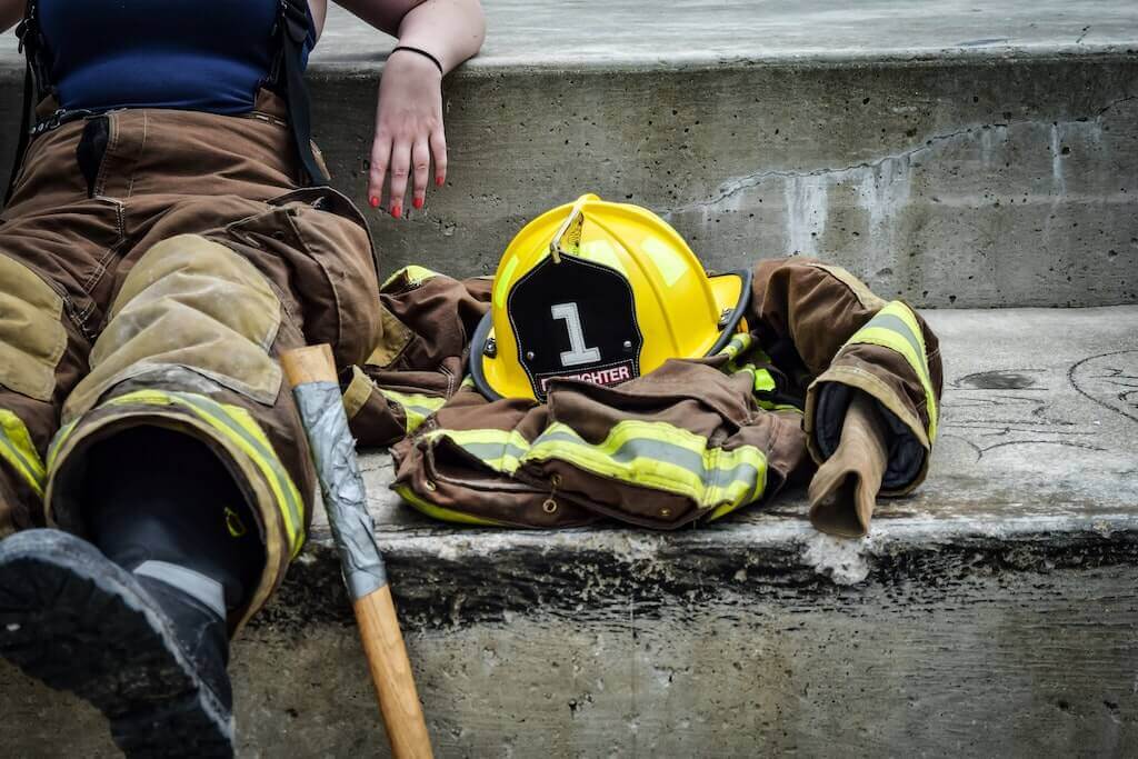 A firefighter sits resting on a concrete step, with turnout gear including a yellow helmet marked '1', a folded protective jacket, and boots laid out beside them, along with an axe handle.