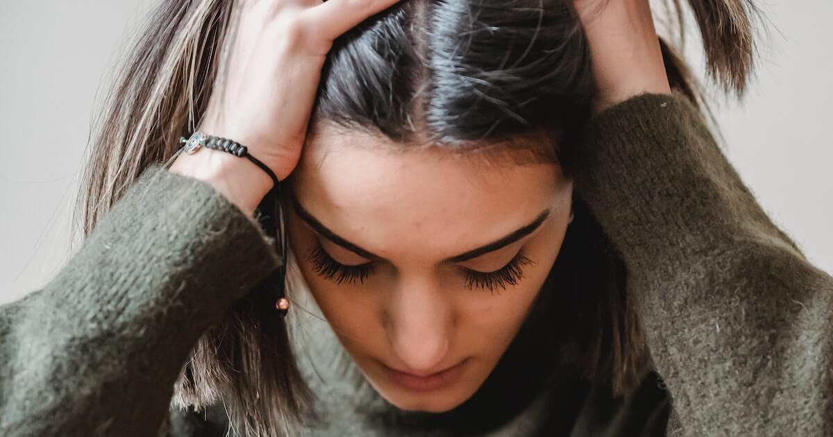 Close-up of a woman with dark hair running her hands through her hair, looking down with a stressed or overwhelmed expression, wearing an olive-green sweater and a bracelet.