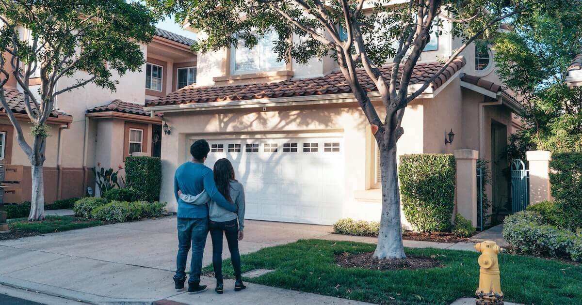 A couple stands arm in arm on a sidewalk, looking up at a two-story stucco house with a tiled roof, garage, and trees in a suburban neighborhood.