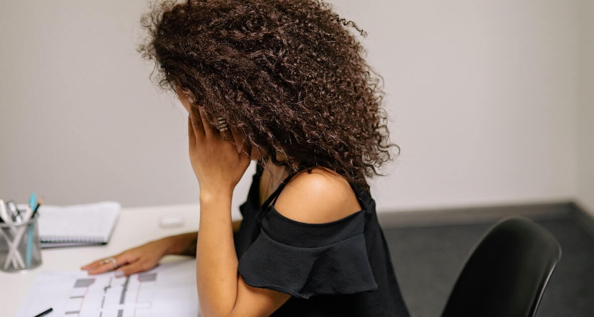 A woman with curly hair sits at a desk with her head resting in her hand, looking down at papers, appearing stressed or tired.