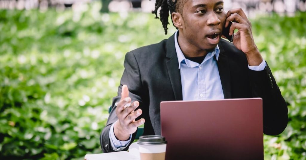 A businessman in a suit sits outdoors talking on his phone with an animated expression, a laptop and coffee cup on the table in front of him, with a green hedge in the background.