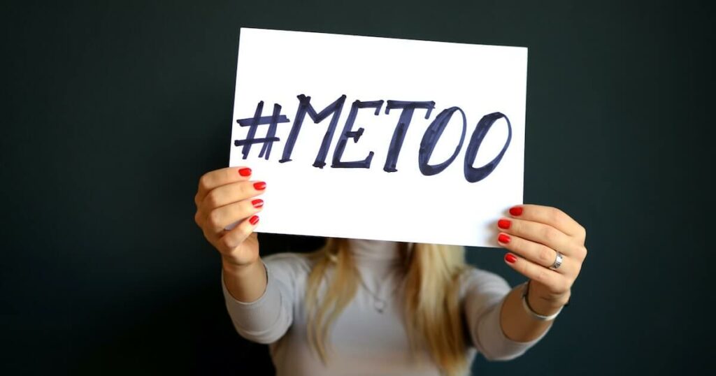 A woman with red nail polish holds up a sign reading '#METOO' in front of her face, standing against a dark background.
