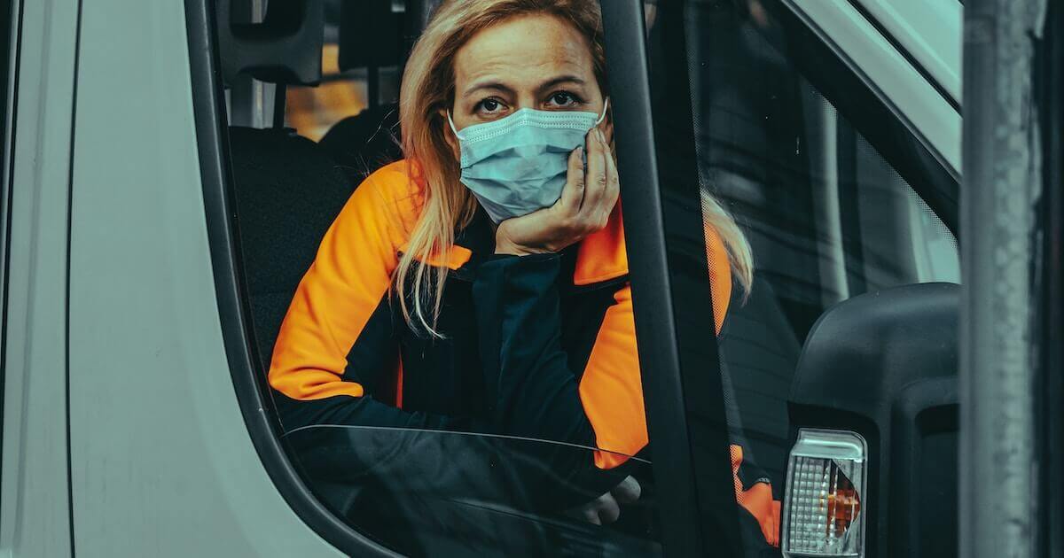 A woman wearing an orange jacket and a face mask looks out from the driver's seat window of a work vehicle.