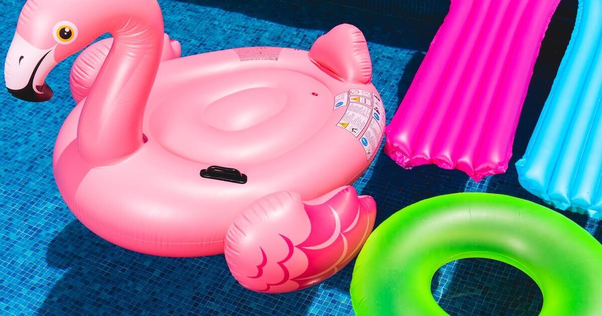Colorful inflatable pool floats — a pink flamingo, a magenta mattress raft, a blue mattress raft, and a green ring float — arranged in a swimming pool.