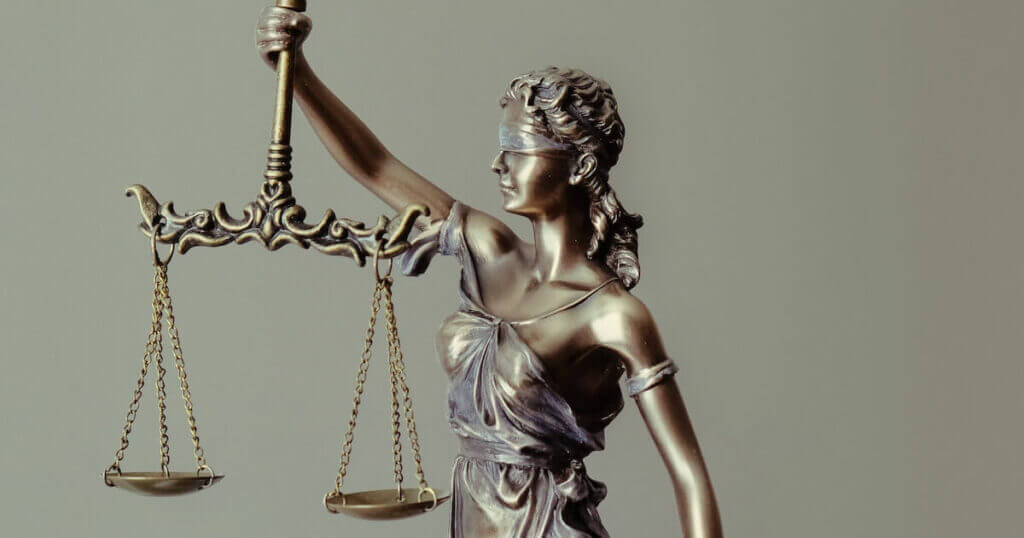 "A bronze statue of Lady Justice, blindfolded and holding a scale, against a plain gray background.