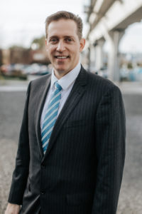 Professional headshot of Thomas L. Spraggs, smiling, wearing a dark pinstripe suit with a light blue striped tie, standing outdoors.